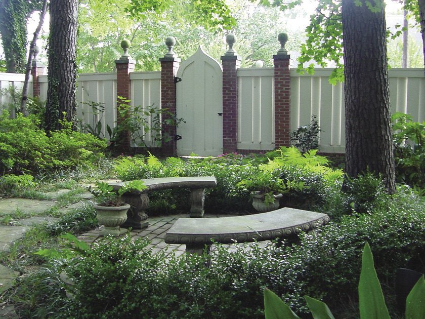 Low green hedges surrounding a circular brick patio (?) with two curved stone benches on either side in front of a white fence with red brick columns.